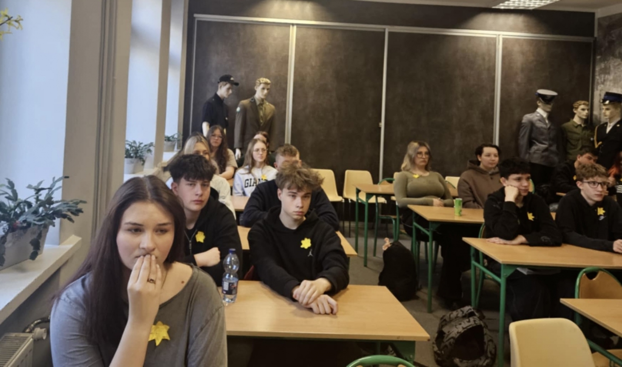 Uczniowie Zespołu Szkół CKZ w Bujnach siedzący w sali lekcyjnej podczas obchodów Akcji Żonkile. Młodzież skupiona, zamyślona, ubrana na ciemno, z przypiętymi papierowymi żonkilami – symbolem pamięci o powstaniu w getcie warszawskim. W tle manekiny w historycznych mundurach, podkreślające edukacyjny charakter spotkania. Na parapetach stoją zielone rośliny, a wystrój sali jest stonowany i poważny, co sprzyja atmosferze zadumy. Uczniowie patrzą uważnie przed siebie, wsłuchani w wykład.