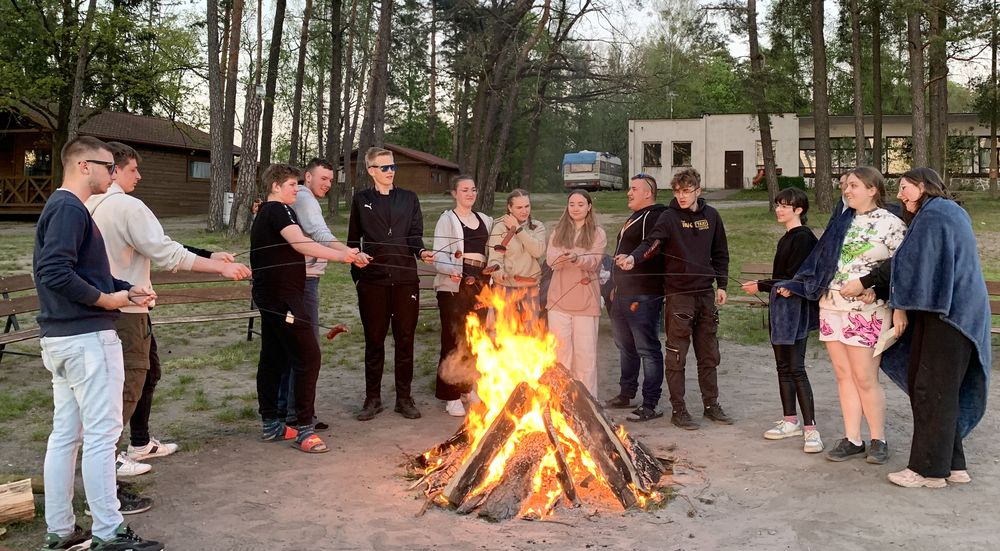 Uczestnicy wycieczki stoją wokół ogniska i pieką kiełbaski