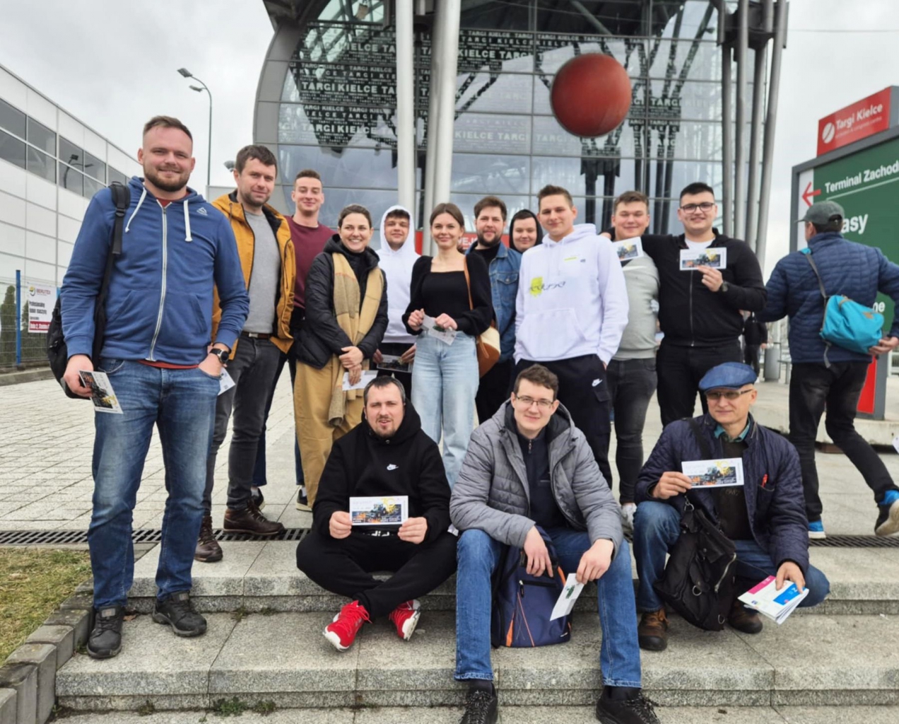 Grupa kilkunastu uczniów i opiekunów z Zespołu Szkół CKZ w Bujnach pozuje przed wejściem do hali wystawienniczej Targi Kielce, gdzie odbywały się XXX Międzynarodowe Targi Techniki Rolniczej Agrotech 2025. W tle widoczna jest nowoczesna szklana fasada budynku z charakterystyczną kulą i nazwą „TARGI KIELCE” umieszczoną na elewacji. Uczestnicy stoją i siedzą na schodach przed wejściem. Uśmiechnięci, trzymają w rękach ulotki i broszury z targów. Młodzież ubrana jest w sportowe kurtki, bluzy i jeansy – styl luźny, dostosowany do wiosennej pogody. Z prawej strony kadru widać duży kierunkowskaz z napisem „Terminal Zachodni” i czerwonym logo targów. Atmosfera na zdjęciu jest swobodna, pełna energii i pozytywnego nastawienia – uczniowie wyglądają na zadowolonych z udziału w wydarzeniu i gotowych do dzielenia się zdobytą wiedzą po powrocie do szkoły.