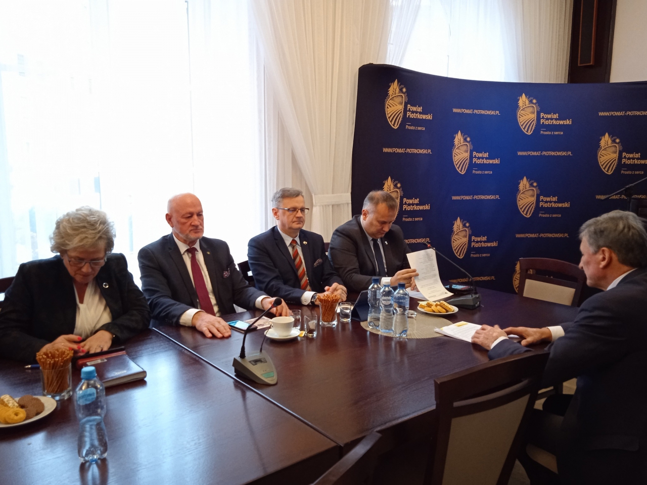 Starosta Piotr Wojtysiak, Wicestarosta Dariusz Magacz oraz Członkowie Zarządu podczas sesji znajdują się w sali konferencyjnej piotrkowskiego Starostwa. w tle napis Powiat Piotrkowski Prosto z serca oraz logo powiatu.