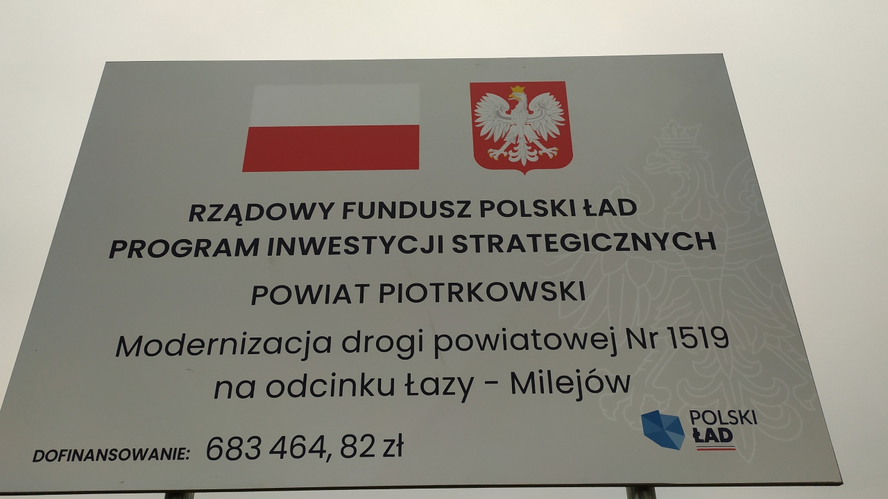 Tablica informacyjna stojąca przy drodze. Rządowy Fundusz Polski Ład. Program Inwestycji Strategicznych. Powiat Piotrkowski. Modernizacja drogi powiatowej Nr 1519E na odcinku Łazy – Milejów. Dofinansowanie 683 464,82 zł. Logo Polskiego Ładu.