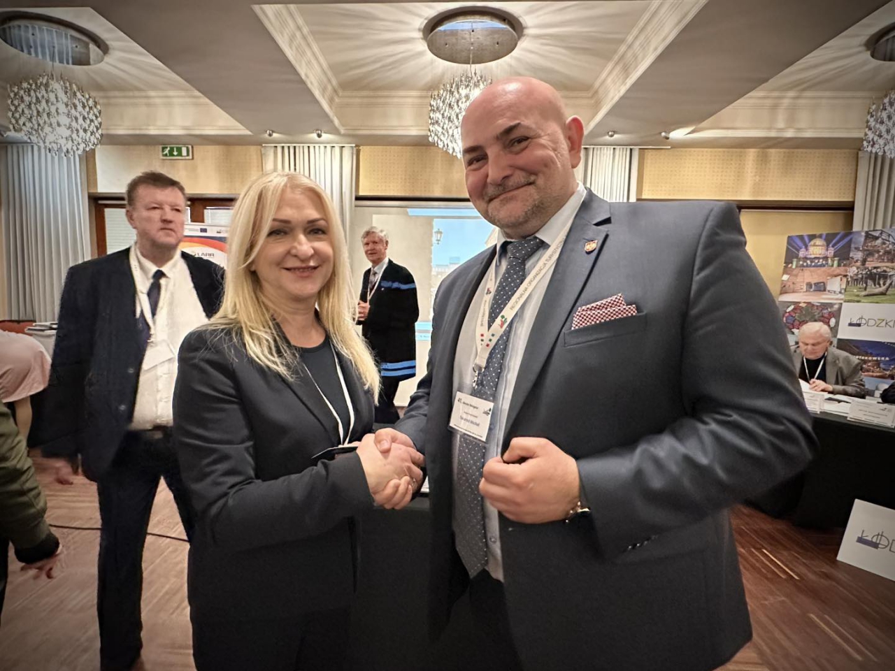 Wicestarosta Powiatu Piotrkowskiego Michał Tokarski ściska dłoń Luizy Staszczak-Gąsiorek, nowo wybranej Prezes Regionalnej Organizacji Turystycznej Województwa Łódzkiego. Oboje uśmiechają się w kierunku aparatu, ubrani elegancko – w tle uczestnicy Walnego Zgromadzenia ROTWŁ oraz materiały promujące województwo łódzkie. Zdjęcie wykonane podczas spotkania w reprezentacyjnej sali konferencyjnej w Łodzi, 26 marca 2025 r.