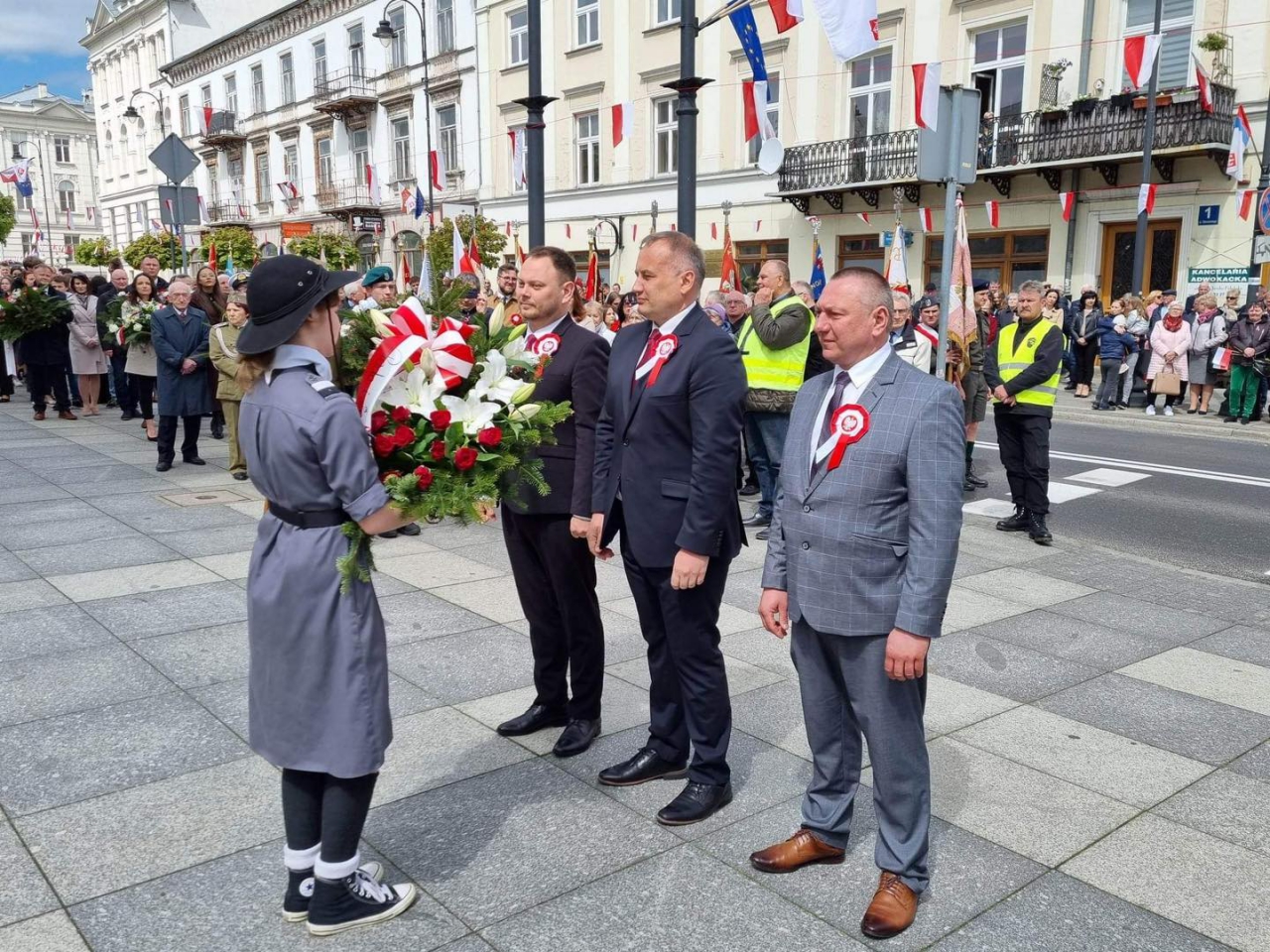 Trzech męźczyzn stoi przed harcerka i jeden z nich przekazuje wiązanke kwiatów, w tle tłum ludzi z biało-czerwonymi flagami