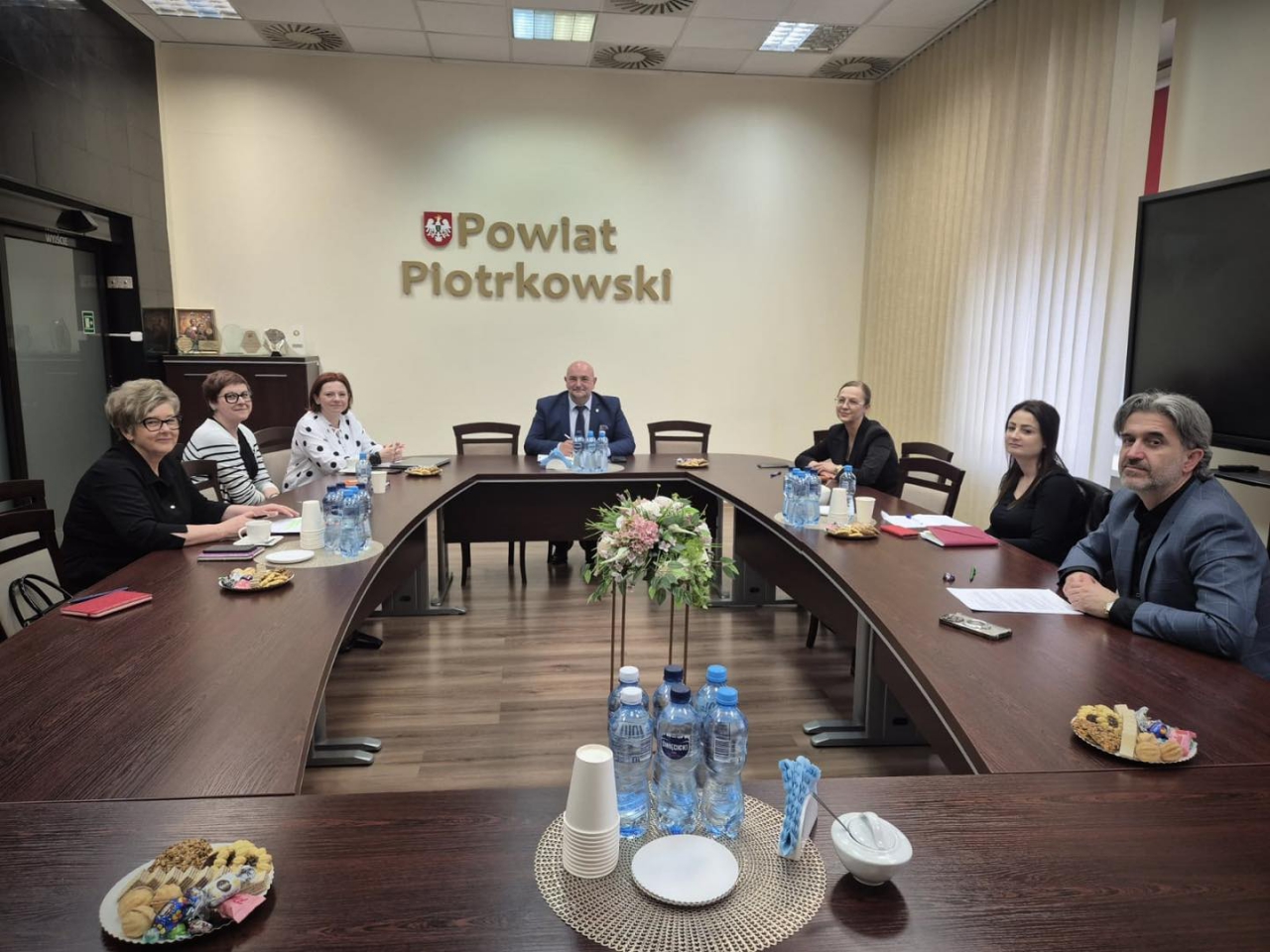 Sala konferencyjna Starostwa Powiatowego w Piotrkowie Trybunalskim. Przy dużym, półokrągłym stole z ciemnego drewna zasiada dziewięcioro uczestników spotkania – przedstawiciele powiatu oraz samorządów gminnych. Na ścianie w tle widoczny jest herb państwowy oraz napis „Powiat Piotrkowski”. Stół udekorowany jest bukietem kwiatów oraz poczęstunkiem: butelkami wody, filiżankami, ciastkami i słodyczami. W tle – jasne rolety i ekran multimedialny. Uczestnicy spotkania uśmiechają się, atmosfera jest życzliwa i robocza.
