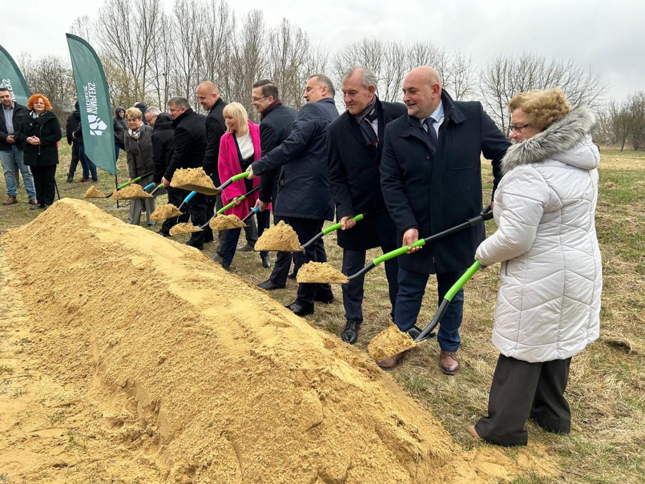 Grupa kilkunastu osób bierze udział w symbolicznym wbiciu łopaty podczas inauguracji budowy Kompleksu Wierzeje. Uczestnicy stoją w szeregu przy pryzmie jasnego piasku, trzymając łopaty z zielonymi trzonkami, nabierają nimi piasek. Wśród obecnych są przedstawiciele samorządu, w tym wicestarosta, oraz osoby zaangażowane w projekt. W tle widać flagi z logo inwestycji i bezlistne drzewa na tle pochmurnego nieba. Wydarzenie ma oficjalny i uroczysty charakter.