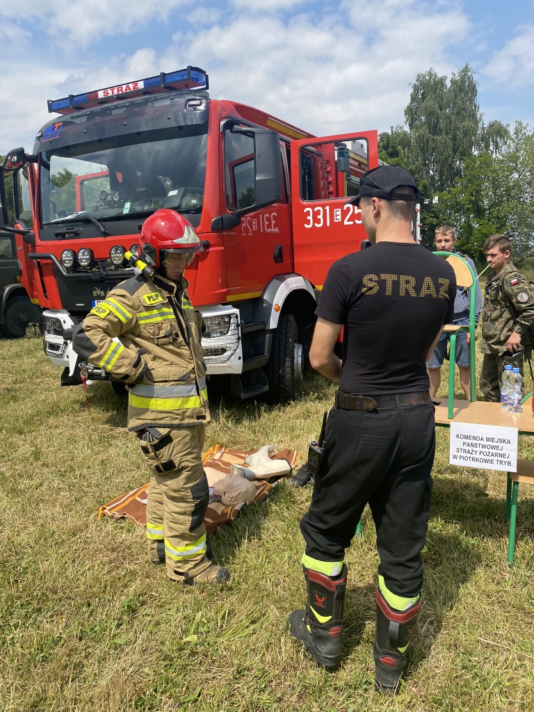 Strażacy podczas pokazu na świezym powietrzu. W tle samochów strażacki.