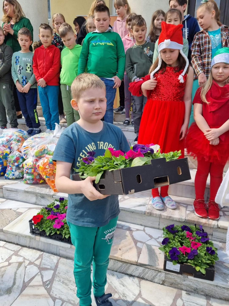Chłopczyk niesie pudełko pełne czerwonych stokrotek, w tle uśmiechają się dzieci, obok których stoja worki pełne kolorowych, plastikowych nakrętek