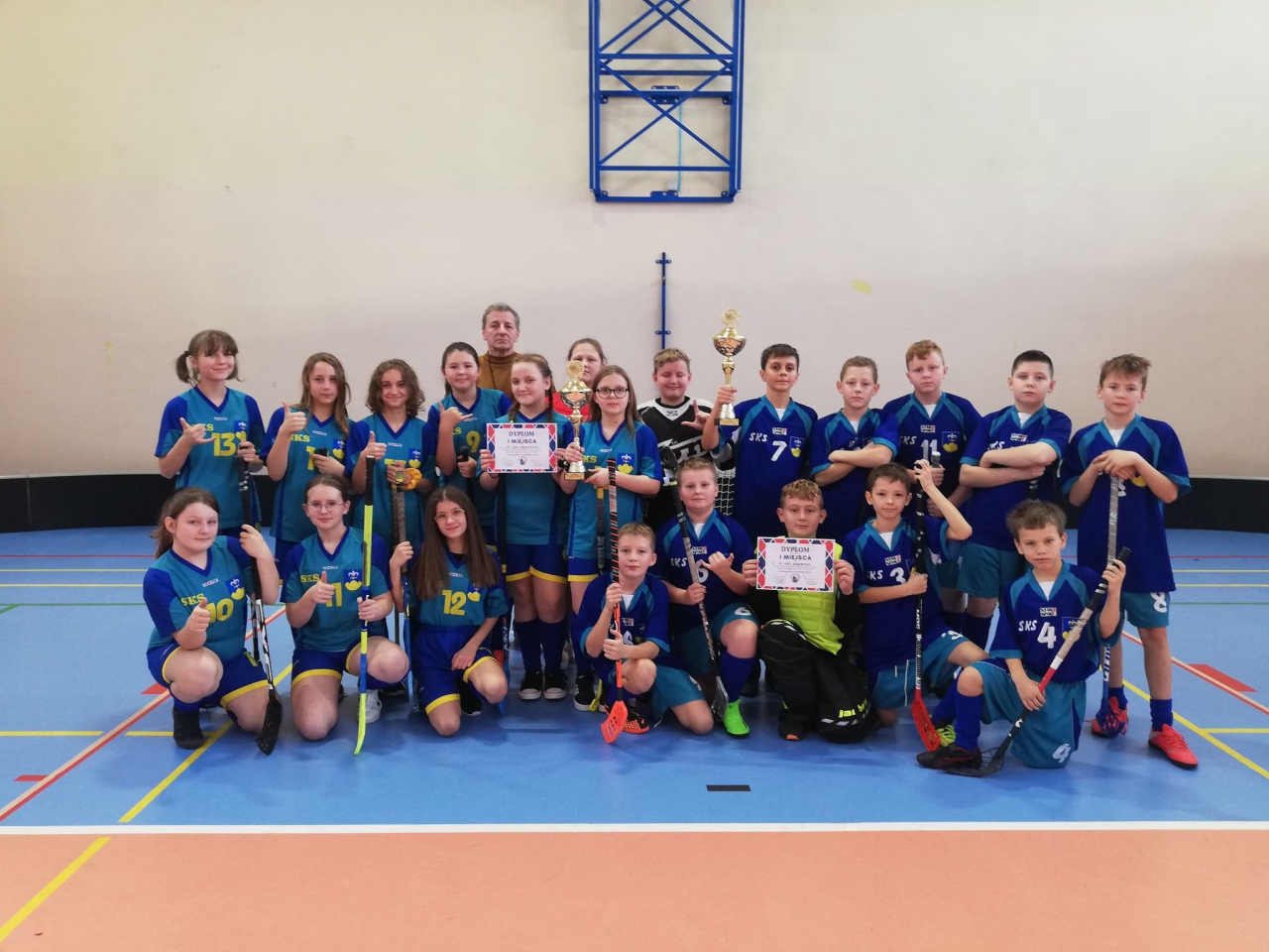 ucznieowe ze szkoły podstawowej w Łękasz Szlacheckich stoja na hali sportowej z pucharami i dyplomami za zdobycie pierwszych miejsc w Mistrzostwach Powiatu Piotrkowskiego w unihokeju