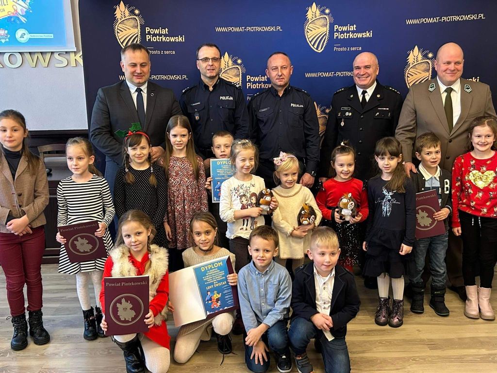 Starosta Piotr Wojtysiak wraz z zaproszonymi gośćmi oraz zwycięzcami konkursu stoją w sali konferencyjnej Starostwa. W tle napis Powiat Piotrkowski oraz logo Powiat prosto z serca.