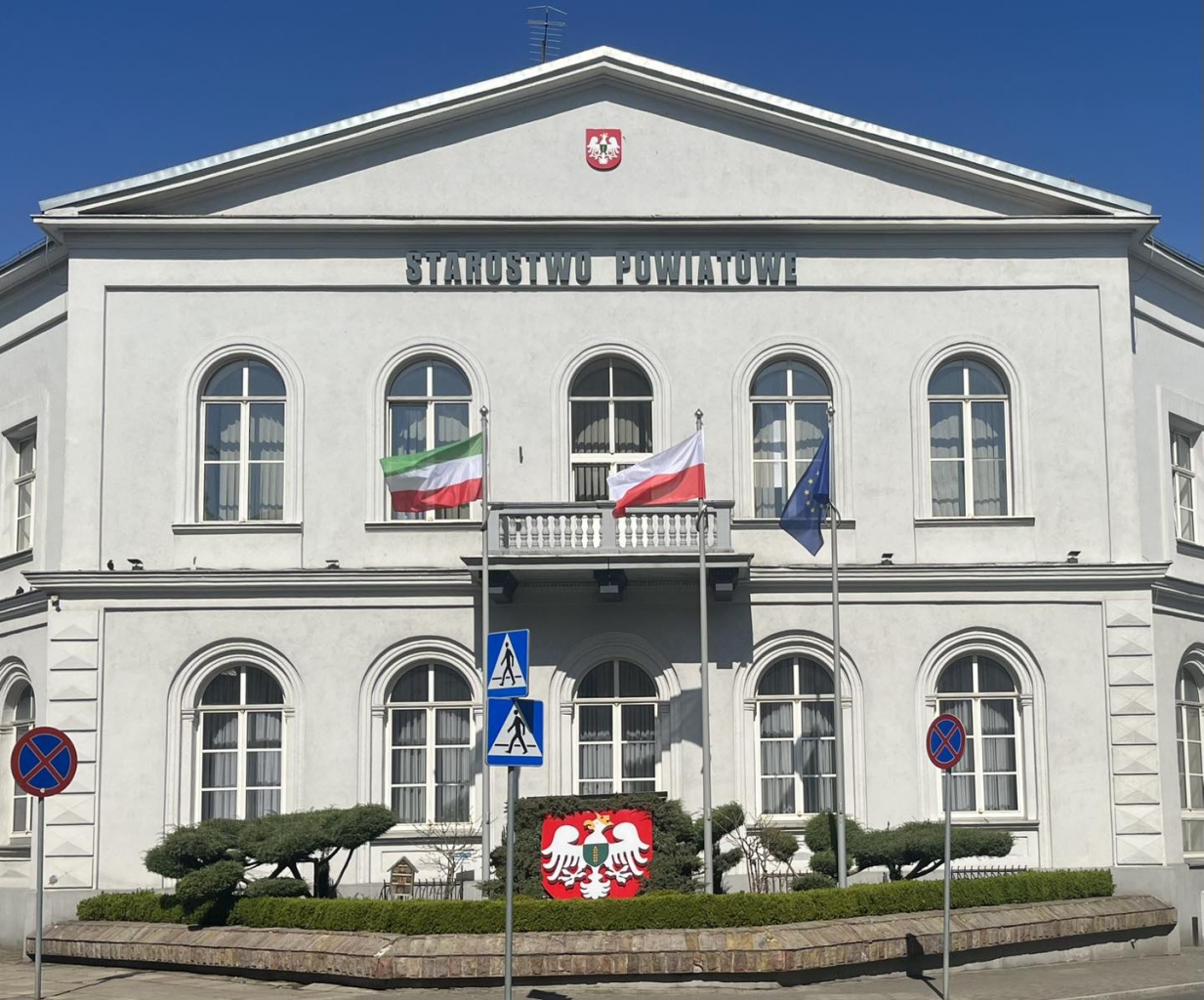 Budynek Starostwa Powiatowego w Piotrkowie Trybunalskim z zewnątrz. Na nim napis starostwo Powiatowe. Przed budynkiem flaga Powiatu w kolorach zielonym, białym, czerwonym, polska flaga biało - czerwona oraz flaga unijna niebieska z żółtymi gwiazdami. Ponadto godło powiaty w kolorach zielonym, białym oraz czerwonym.