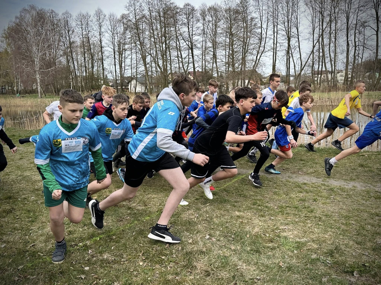 Grupa chłopców w różnokolorowych strojach sportowych rusza dynamicznie ze startu podczas biegu przełajowego. W tle widoczne są drzewa bez liści i fragment stawu. Zawodnicy biegną po trawiastej ścieżce, skupieni i pełni energii. To moment rozpoczęcia rywalizacji podczas Mistrzostw Powiatu Piotrkowskiego w drużynowych biegach przełajowych.