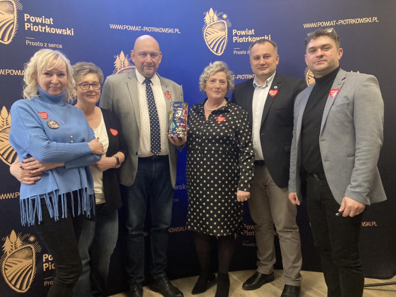 Starosta Piotr Wojtysiak wraz z pozostałymi władzami Powiatu, naczelnym kierownictwem i Radnymi znajdują się w sali konferencyjnej Starostwa. Członek zarządu trzyma puszkę WOŚP. W tle napis i logo Powiat Piotrkowski Prosto z serca.