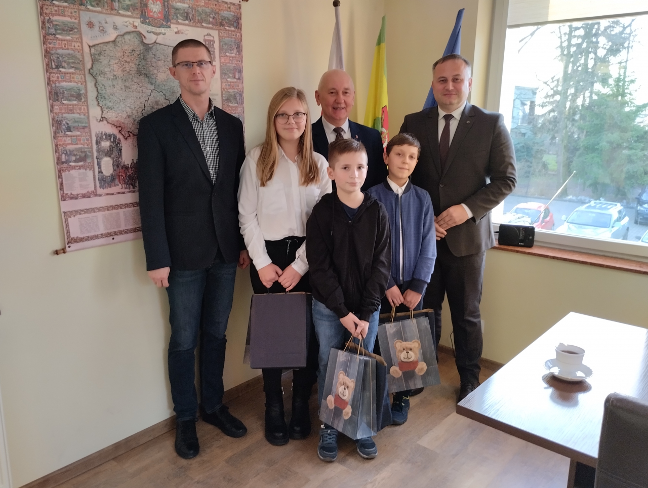 Starosta Piotr Wojtysiak oraz Wójt Gminy Moszczenica Marceli Piekarek wraz z Laureatmi Konkursu stoją w Urzędzie Gminy w Moszczenicy. W tle historyczna mapa Polski.