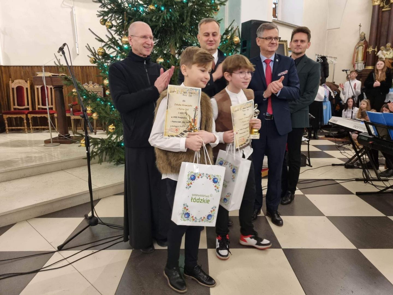 Starosta Piotr Wojtysiak oraz Wicestarosta Dariusz Magacz stoją z Laureatami Konkursu w Kościele w Moszczenicy. W tle choinka.