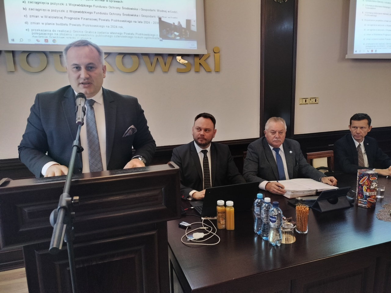 Starosta przemawia z mównicy, obok siedzi prezydium Rady Powiatu