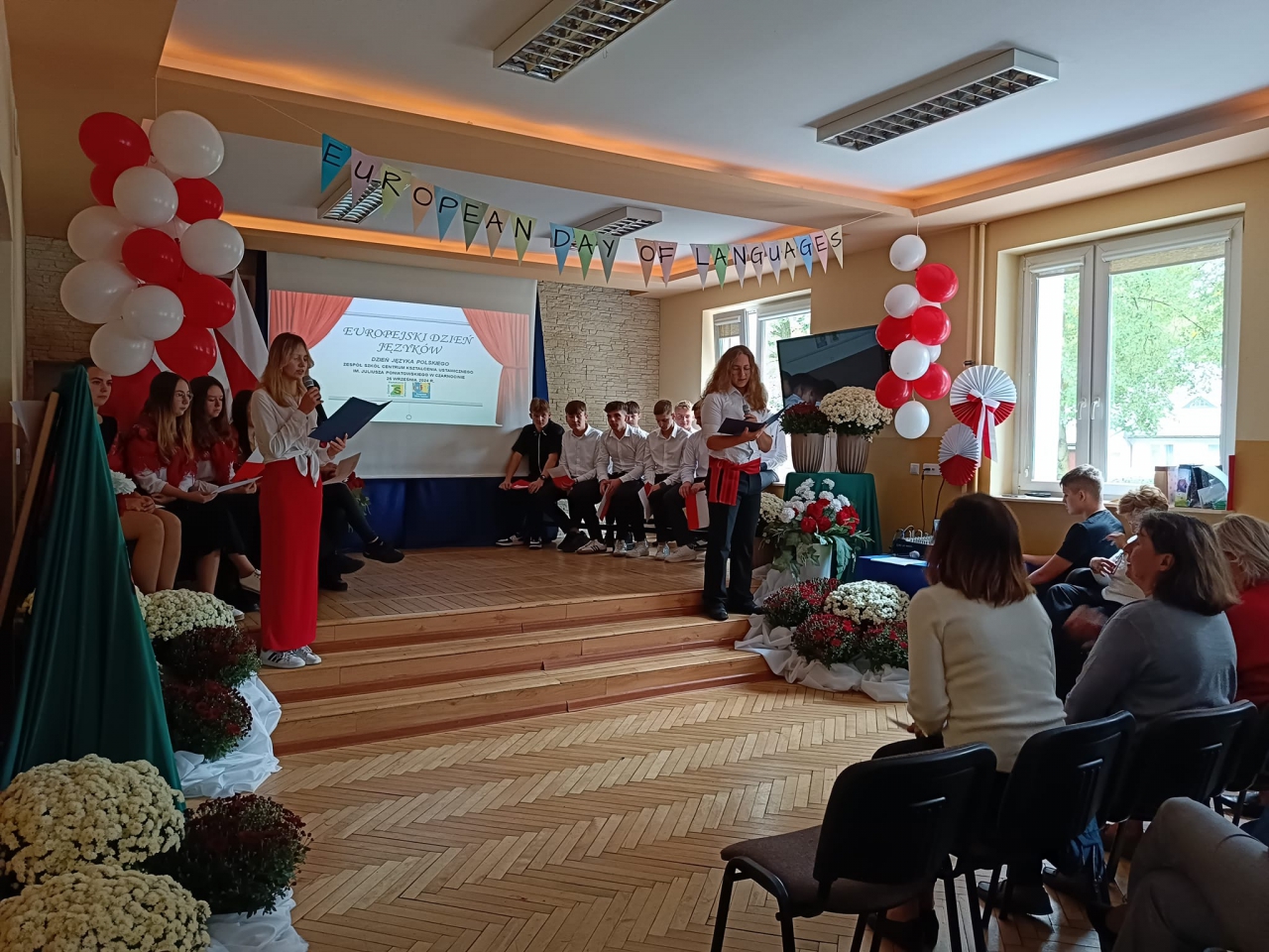 Dziewczyna i chłopak stoją po bokach z mikrofonem i kartką w rękach. Na środku napis Europejski Dzień Języków Obcych, w tle balony białoczerwone, kwiaty inne osoby