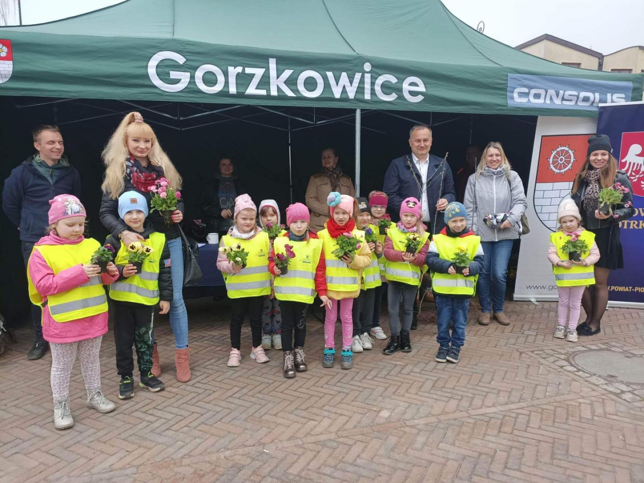 Na zdjęciu stoją dzieci z kwiatkami i osoby dorosłe