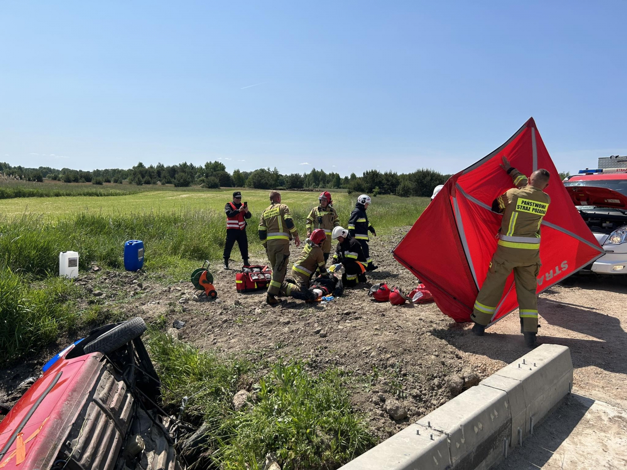 Strażacy udzielają pomocy rannemu, w rzece leży przewrócone auto