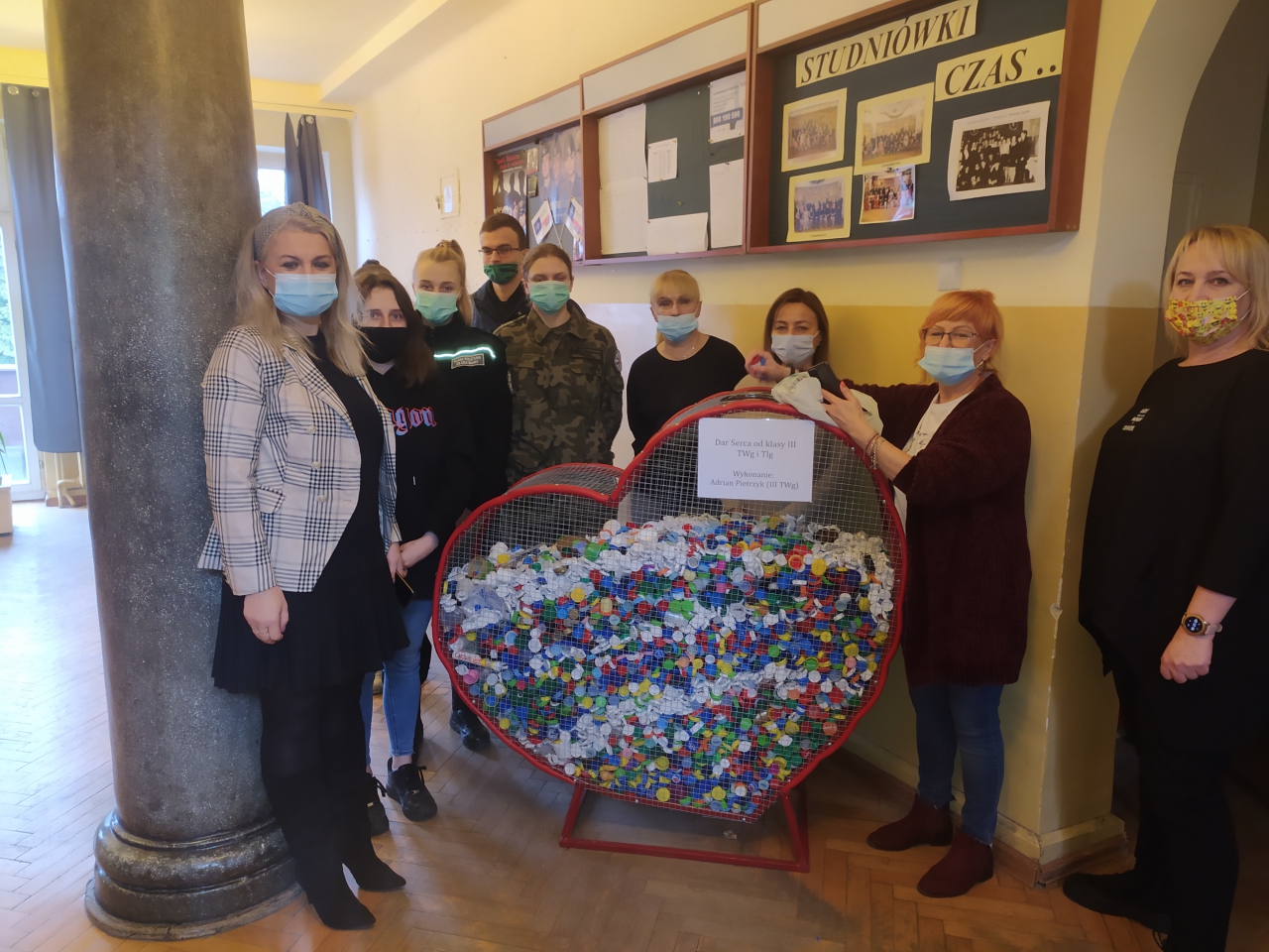 Młodzież wraz z dyrekcją szkoły stoi przy metalowym sercu wypełnionym nakrętkami