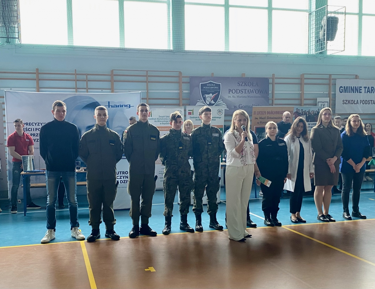 Dyrekcja szkoły w Bujanch oraz uczniowie stoją w pomieszczeniu podczas targów w Gomulinie. W tle nośniki wystawiennicze z nazwą szkoły oraz logo szkoły.