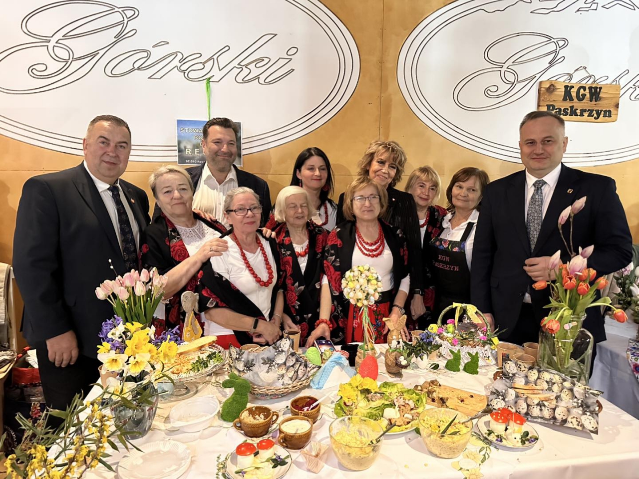 Grupowe zdjęcie przy stoisku Koła Gospodyń Wiejskich w Paskrzynie podczas Jarmarku Wielkanocnego. Uczestnicy stoją za bogato udekorowanym stołem pełnym tradycyjnych potraw, pisanek i kwiatów. Wśród nich widoczni są członkowie KGW w regionalnych strojach z czerwonymi akcentami oraz przedstawiciele władz samorządowych. W tle duże logo „Górski” oraz tabliczka z napisem „KGW Paskrzyn”.