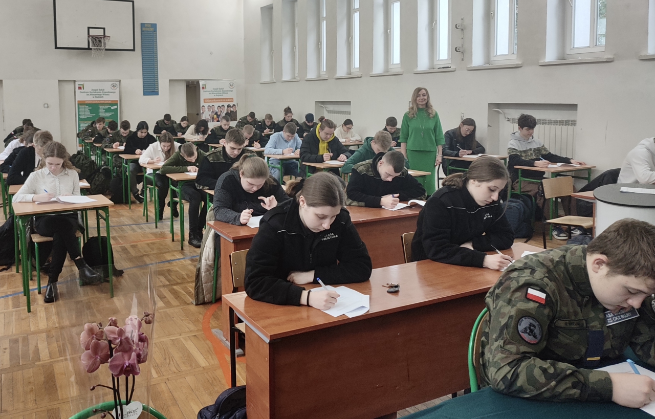 sala gimnastyczna uczniowie siedząc przy ławkach rozwiązują test