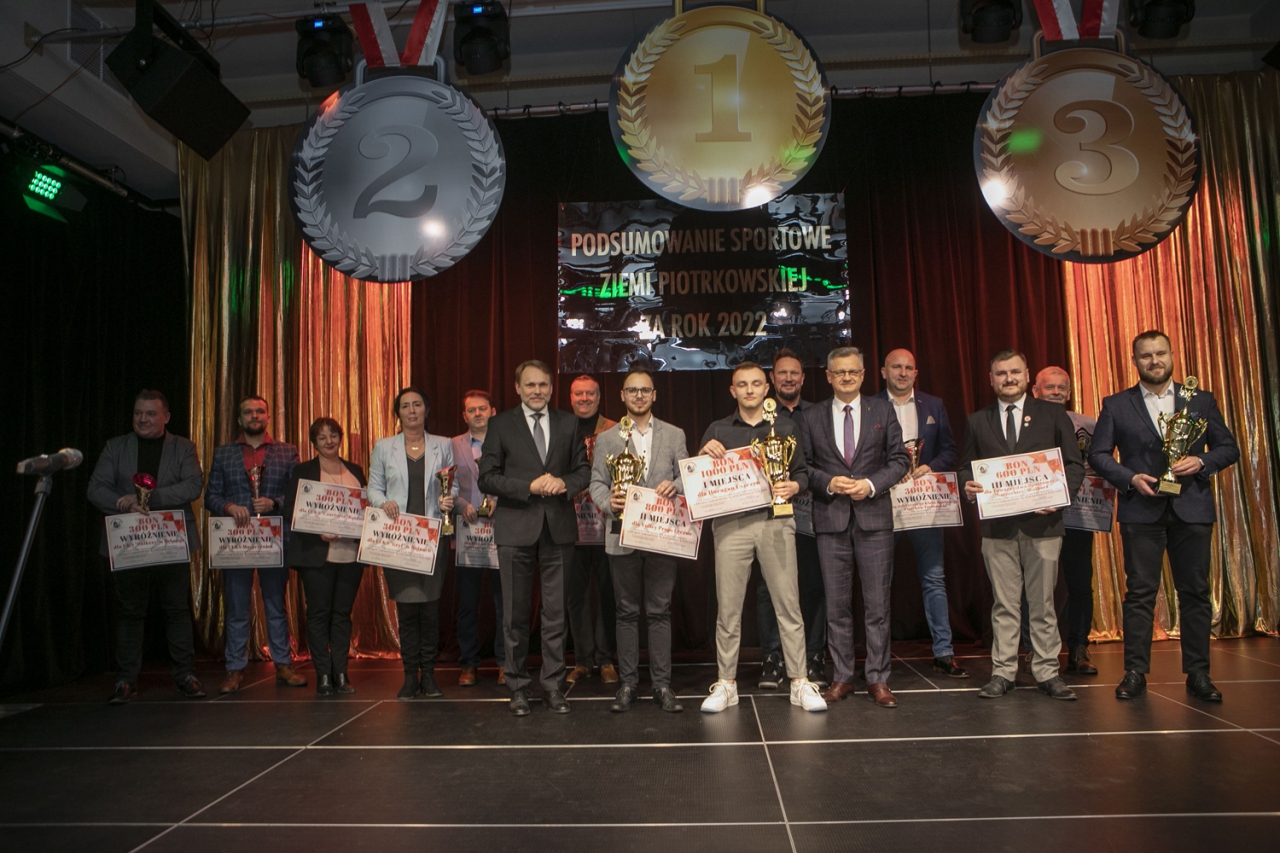 Władze Powiatu oraz zaproszeni goście stoją z nagrodzonymi na scenie w sali w Grabicy. Nad sceną wiszą trzy medale a w tle napis Podsumowanie Sportowe Ziemi Piotrkowskiej za 2022 rok.