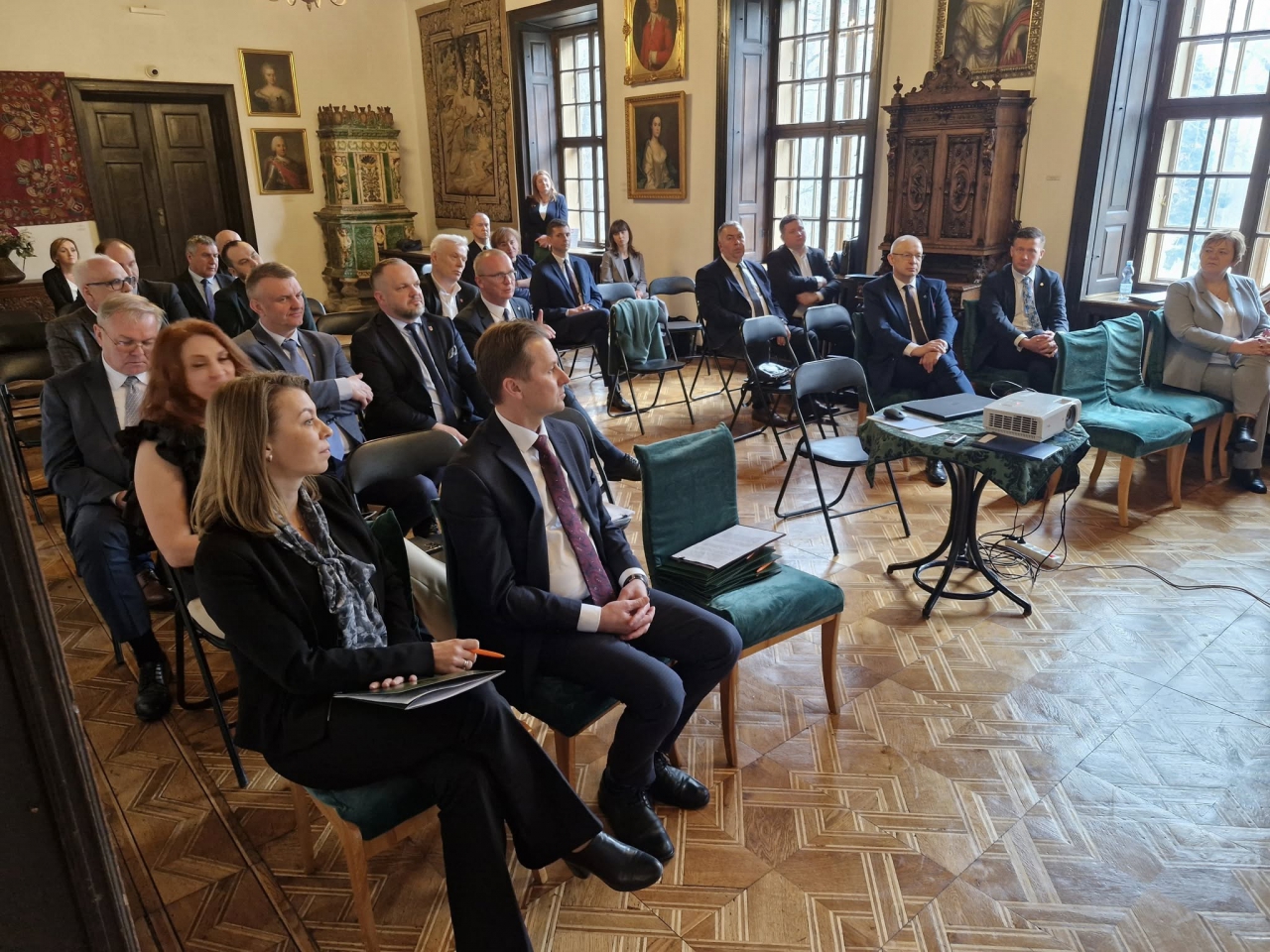 Sala Konwentu Starostów w Oporowie, w zabytkowym wnętrzu pałacowym. Uczestnicy wydarzenia – starostowie i zaproszeni goście – siedzą w skupieniu na zielonych i czarnych krzesłach ustawionych w kilku rzędach. W tle widoczne są stylizowane piece kaflowe, drewniane meble oraz portrety historyczne zawieszone na ścianach. Pomieszczenie ma duże, drewniane okna, przez które wpada naturalne światło. Na pierwszym planie widoczny jest projektor ustawiony na ozdobnym stoliku z zielonym obrusem, a obok leżą przewodniki lub materiały konferencyjne. Atmosfera jest poważna i uroczysta, a uczestnicy uważnie śledzą przebieg spotkania.