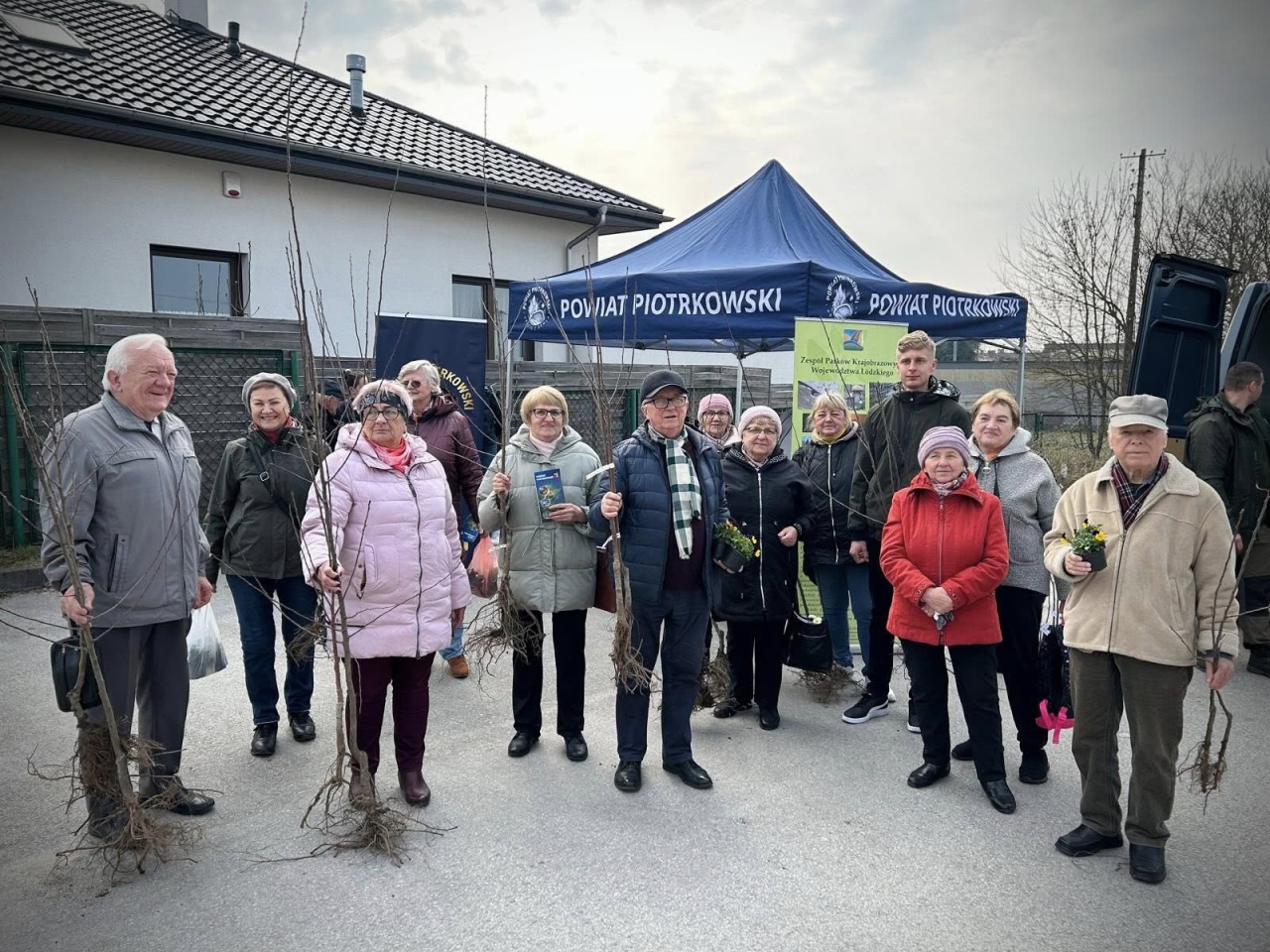 Grupa uczestników akcji „Nakręć się na zieleń” pozuje do wspólnego zdjęcia przy namiocie Powiatu Piotrkowskiego. W tle widoczny baner Zespołu Parków Krajobrazowych Województwa Łódzkiego. Uśmiechnięci mieszkańcy, głównie seniorzy, trzymają w rękach sadzonki drzewek i doniczki z kwiatami. Zdjęcie wykonane na świeżym powietrzu, przy zabudowaniach jednorodzinnych, w pogodny, choć lekko pochmurny dzień.