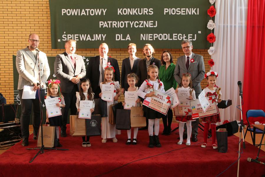 Starosta Piotr Wojtysiak oraz Wicestarosta Dariusz Magacz stoją na scenie w sali gimnastycznej w Łękach Szlacheckich wraz ze zwycięzcami konkursu oraz członkami jury. W tle napis Powiatowy Konkurs Piosenki Patriotycznej Śpiewajmy dla Niepodległej.