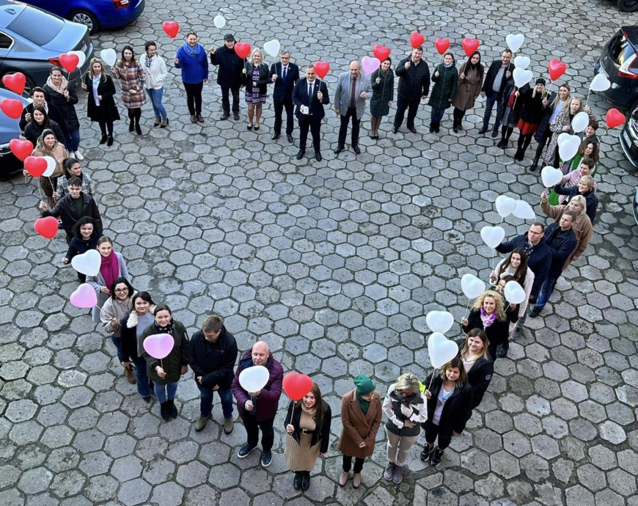  Władze oraz pracownicy Starostwa stoją, ustawieni na kształt serca, na dziedzińcu Urzędu z kolorowymi balonami w rękach.
