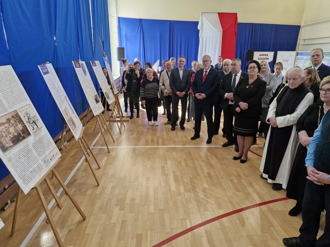 Grupa uczestników wydarzenia zgromadzona wokół wystawy historycznej upamiętniającej 120. rocznicę strajków szkolnych 1905 roku. Na pierwszym planie znajdują się plansze z opisami i archiwalnymi fotografiami, ustawione na drewnianych sztalugach. W tle widać patriotyczną dekorację w barwach biało-czerwonych. Wśród zgromadzonych osób są przedstawiciele władz samorządowych, duchowieństwo oraz zaproszeni goście, którzy z uwagą śledzą treść prezentowanych materiałów