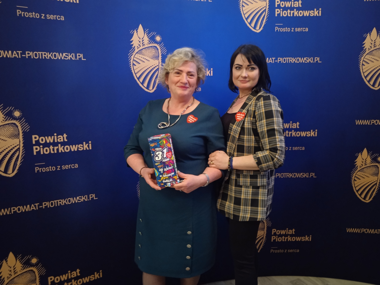 Członek zarządu trzyma puszkę WOŚP. Obok w sali konferencyjnej Starostwa stoi pracownik Starostwa. W tle napis i logo Powiat Piotrkowski Prosto z serca.