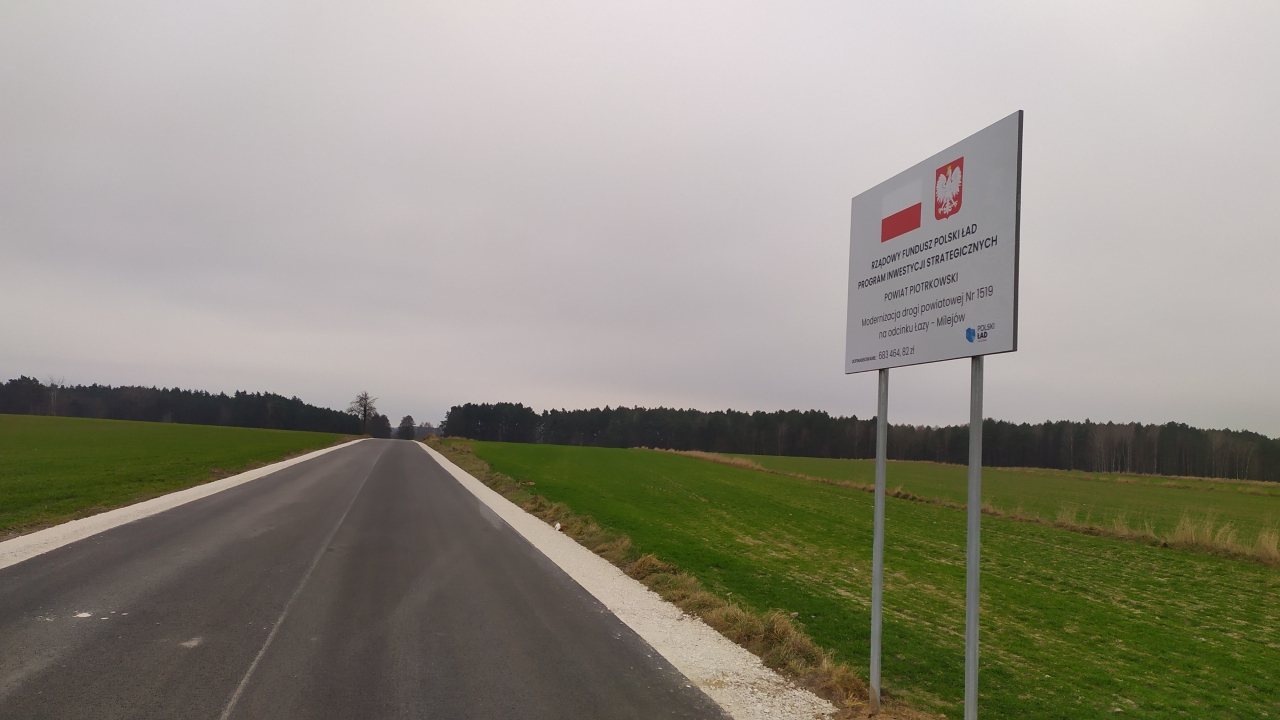Fragment drogi powiatowej Łazy - Milejów. Po prawej stronie tablica informująca o otrzymanym dofinansowaniu.