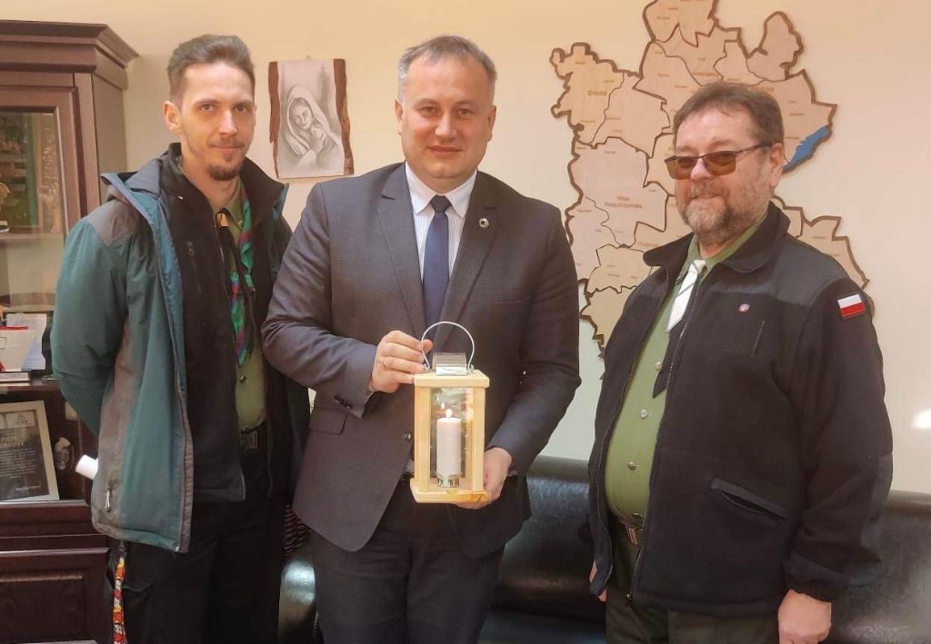Starosta Piotr Wojtysiak wraz z przedstawicielami piotrkowskiego Hufca stoi w pomieszczeniu z betlejemskim światełkiem. W tle mapa Powiatu Piotrkowskiego.