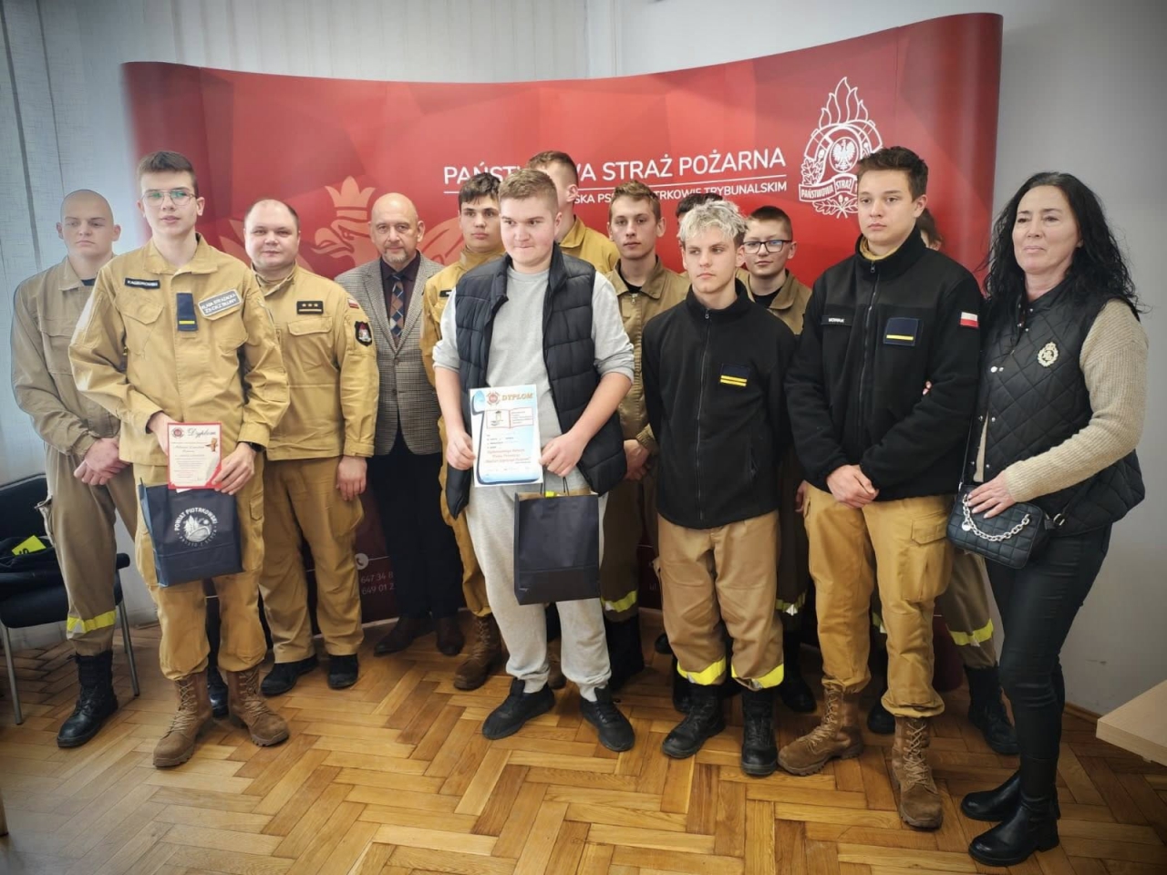 Grupa uczestników konkursu wiedzy pożarniczej pozuje do wspólnego zdjęcia z opiekunami i przedstawicielem Powiatu Piotrkowskiego na tle ścianki Komendy Miejskiej PSP w Piotrkowie Trybunalskim. Uczniowie trzymają dyplomy i nagrody, uśmiechnięci i dumni z udziału w wydarzeniu.