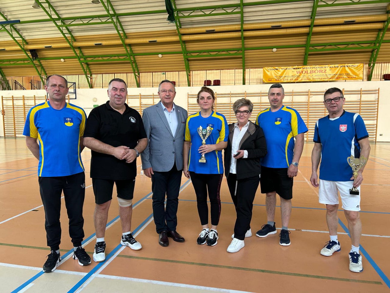 Grupa siedmiorga dorosłych osób pozuje wspólnie na tle hali sportowej, w&nbsp;której odbyła się XXIV Spartakiada Władz Samorządowych w&nbsp;Badmintona. W&nbsp;tle widoczne są wysokie okna, jasne ściany z&nbsp;drabinkami gimnastycznymi i&nbsp;baner informujący o&nbsp;lokalnym klubie sportowym w&nbsp;Wolborzu.  Na zdjęciu, od lewej: – Mężczyzna w&nbsp;niebiesko-żółtej koszulce z&nbsp;herbem gminy Wolbórz i&nbsp;czarnych spodniach dresowych, – Mężczyzna w&nbsp;czarnej koszulce i&nbsp;czarnych spodenkach, z&nbsp;opaską na kolanie, – Mężczyzna w&nbsp;jasnoszarej marynarce, granatowych spodniach i&nbsp;brązowych butach, – Kobieta w&nbsp;niebiesko-żółtej koszulce, trzymająca puchar, – Kobieta w&nbsp;czarnej kurtce i&nbsp;białej koszuli, trzymająca telefon, – Mężczyzna w&nbsp;niebiesko-żółtej koszulce z&nbsp;herbem gminy Wolbórz, – Mężczyzna w&nbsp;niebieskiej koszulce z&nbsp;herbem Powiatu Piotrkowskiego i&nbsp;białych spodenkach, trzymający puchar.  Wszyscy uśmiechnięci, ustawieni w&nbsp;rzędzie na środku boiska, celebrują wspólny sukces i&nbsp;zakończenie wydarzenia sportowego.