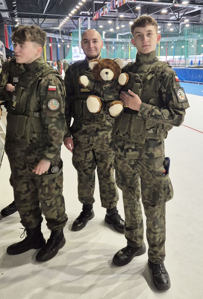 Na zdjęciu trzech młodych uczniów z&nbsp;klasy Oddziału Przygotowania Wojskowego ZS CKZ w&nbsp;Bujnach pozuje w&nbsp;mundurach wojskowych wewnątrz Areny Lodowej w&nbsp;Tomaszowie Mazowieckim. Dwóch z&nbsp;nich stoi przodem do obiektywu, trzeci delikatnie odwrócony w&nbsp;bok. W&nbsp;centralnym punkcie fotografii jeden z&nbsp;uczniów trzyma pluszowego misia ubranego w&nbsp;mundur, co dodaje zdjęciu lekkiego i&nbsp;przyjaznego charakteru. Tło stanowi hala sportowa z&nbsp;widocznymi flagami, oświetleniem i&nbsp;uczestnikami wydarzenia.